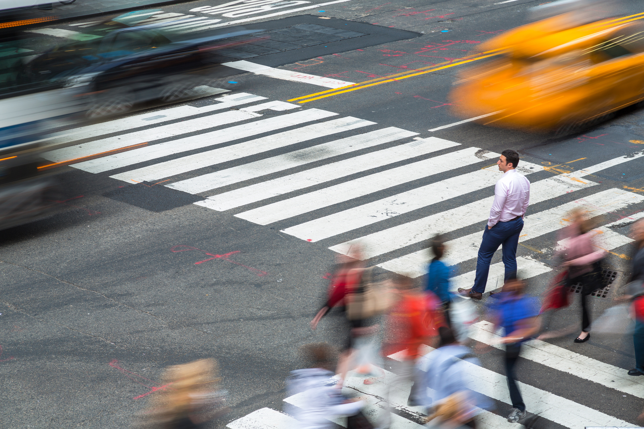 A man pauses at a city crossing as pedestrians and yellow taxis blur past