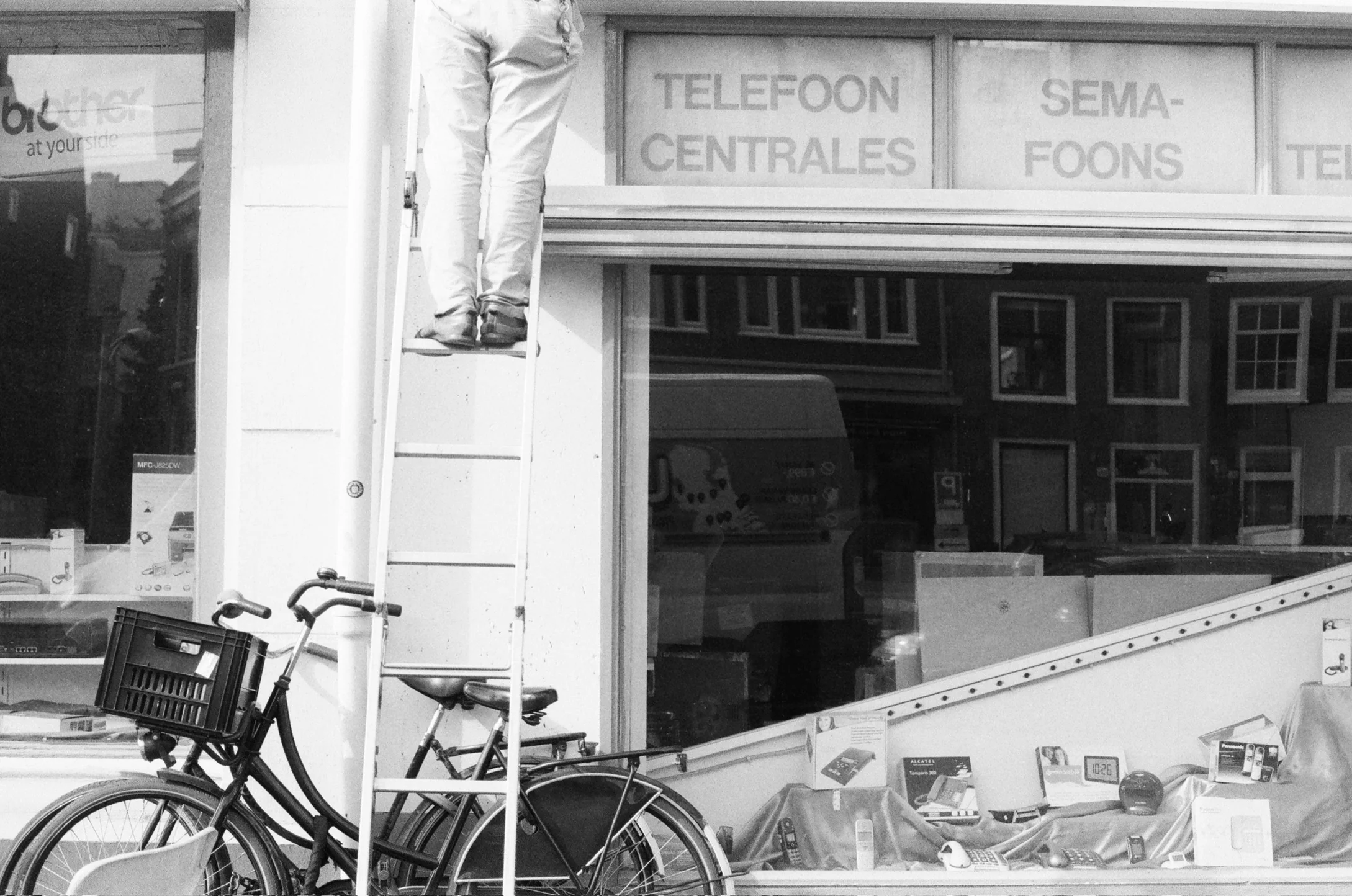 A black-and-white street photograph of a worker on a ladder above bicycles outside a telephone shop