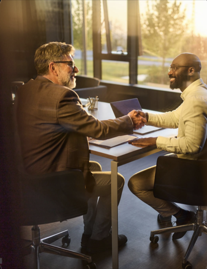 Two men shaking hands across a desk in a warmly sunlit office